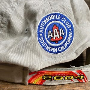 May include: A light grey baseball cap featuring the "AAA Automobile Club Southern California" logo. The peak has "2004" embroidered in red, yellow, and orange. The cap is made of a sturdy fabric and is designed for sun protection.