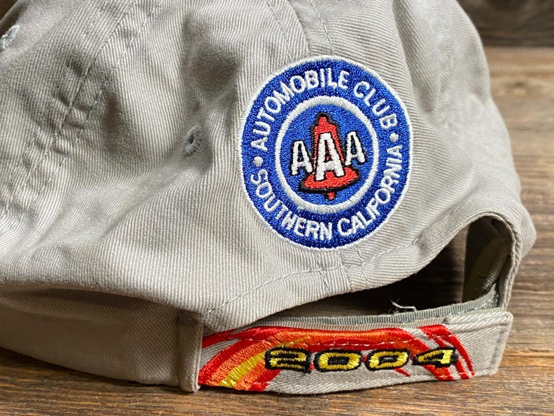 May include: A light grey baseball cap featuring the "AAA Automobile Club Southern California" logo. The peak has "2004" embroidered in red, yellow, and orange. The cap is made of a sturdy fabric and is designed for sun protection.