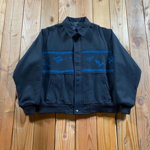 May include: A black jacket featuring a horizontal band across the chest with blue rope-like borders and blue symbols. The jacket has a standard collar, long sleeves with ribbed cuffs, a ribbed hem, and a front button closure with a hidden placket.
