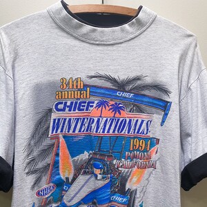 May include: Gray t-shirt with a black mock neck. The front of the shirt features a colorful graphic of a race car with flames and palm trees. The text "34th annual CHIEF WINTERNATIONALS 1994 Pomona California" is printed on the shirt.