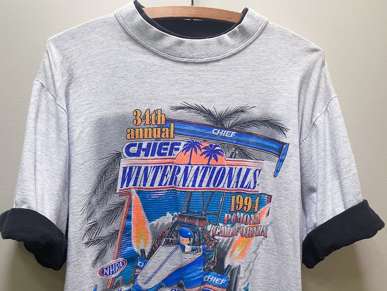May include: Gray t-shirt with a black mock neck. The front of the shirt features a colorful graphic of a race car with flames and palm trees. The text "34th annual CHIEF WINTERNATIONALS 1994 Pomona California" is printed on the shirt.