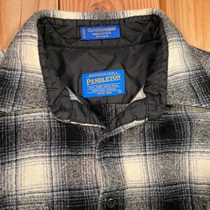 May include: A close-up of a black, white, and grey plaid shirt. The shirt features a black quilted collar and two blue tags with white text. One tag reads "OUTDOORSMAN MADE IN U.S.A." and the other reads "PENDLETON 100% PURE WOOL".