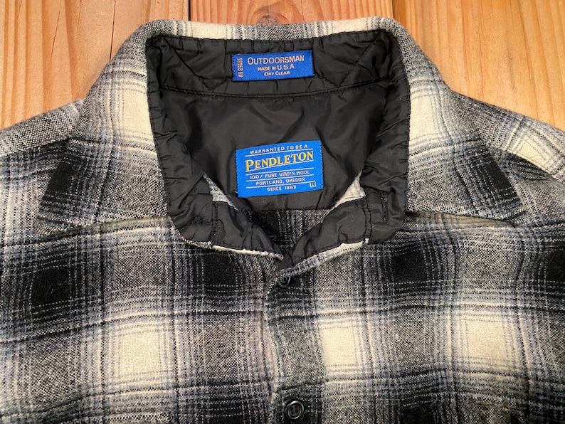 May include: A close-up of a black, white, and grey plaid shirt. The shirt features a black quilted collar and two blue tags with white text. One tag reads "OUTDOORSMAN MADE IN U.S.A." and the other reads "PENDLETON 100% PURE WOOL".