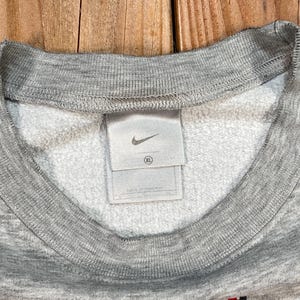 Puede incluir: Primer plano de una sudadera gris con interior blanco. El cuello y los pu&ntilde;os son acanalados. Se ve una etiqueta blanca con el logotipo negro de Nike. La etiqueta indica la talla XL.