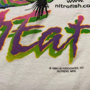 Puede incluir: Camiseta blanca con la palabra "HEAT" en letras grandes, estilizadas y multicolores. El texto est&aacute; delineado en verde y relleno con degradados morados y rosas. La direcci&oacute;n del sitio web "nitrofish.com" es visible.
