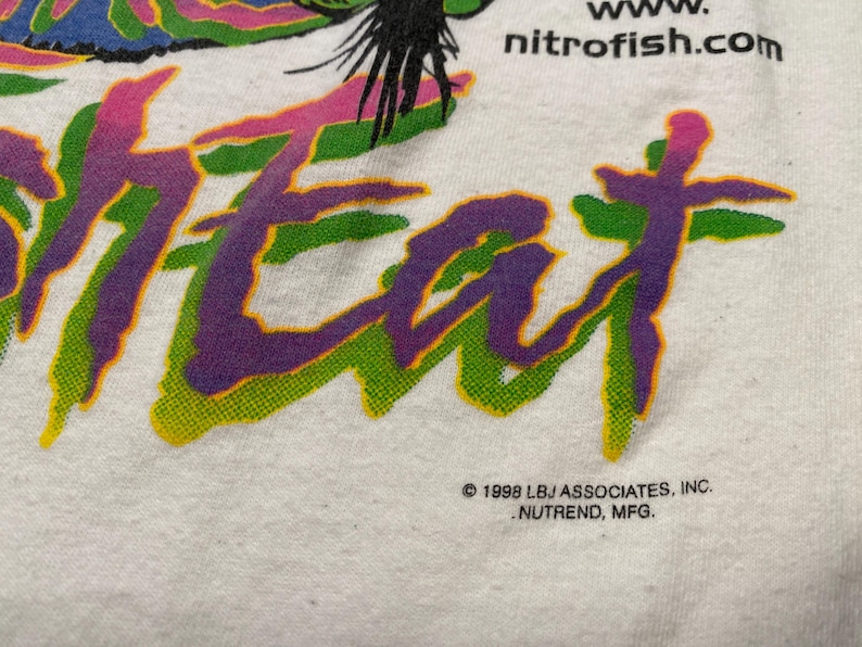 Puede incluir: Camiseta blanca con la palabra "HEAT" en letras grandes, estilizadas y multicolores. El texto est&aacute; delineado en verde y relleno con degradados morados y rosas. La direcci&oacute;n del sitio web "nitrofish.com" es visible.