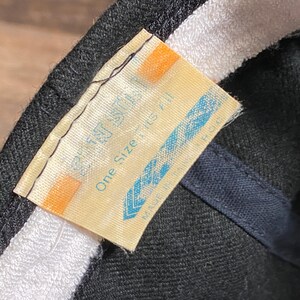 May include: Close-up of a black baseball cap with a white interior band. A tag with the text "One Size Fits All" is visible. The tag is light yellow with blue and orange accents.