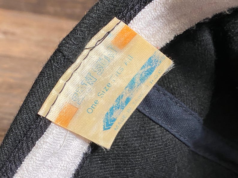 May include: Close-up of a black baseball cap with a white interior band. A tag with the text "One Size Fits All" is visible. The tag is light yellow with blue and orange accents.