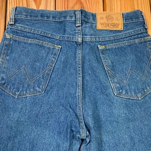May include: Close-up of the back of blue denim jeans. The jeans have two back pockets with a signature W-shaped stitching design. A brown leather patch with the "Wrangler" logo is visible at the waistband. The jeans are made of a medium-wash denim.