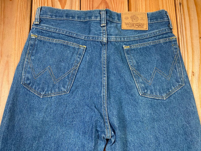 May include: Close-up of the back of blue denim jeans. The jeans have two back pockets with a signature W-shaped stitching design. A brown leather patch with the "Wrangler" logo is visible at the waistband. The jeans are made of a medium-wash denim.