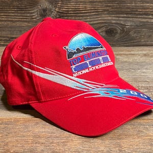 Op de afbeelding: Een felrode honkbalpet met een geborduurd logo met de tekst TOP ELIMINATOR CLUB NATIONAL HOT ROD ASSOCIATION en een blauwe afbeelding van een raceauto. De pet heeft witte en blauwe golvende lijnen en het jaartal 2003 op de klep.