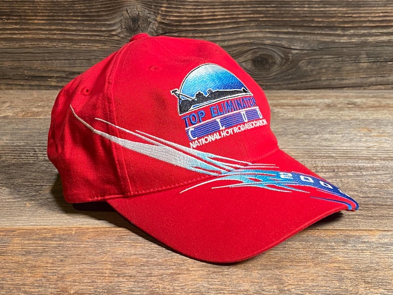 Op de afbeelding: Een felrode honkbalpet met een geborduurd logo met de tekst TOP ELIMINATOR CLUB NATIONAL HOT ROD ASSOCIATION en een blauwe afbeelding van een raceauto. De pet heeft witte en blauwe golvende lijnen en het jaartal 2003 op de klep.