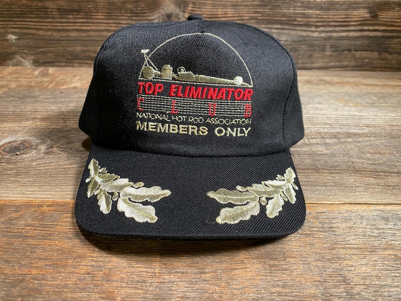 May include: Black baseball cap with embroidered text "TOP ELIMINATOR" and "MEMBERS ONLY". Features a racing car graphic and leaf designs on the brim. The cap is made of a dark fabric.