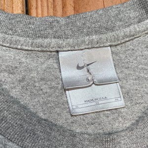 Puede incluir: Primer plano de un cuello de camiseta gris con una etiqueta Nike. La etiqueta es blanca con un logotipo Nike gris y el texto "MADE IN U.S.A.". La camiseta es de color gris liso.