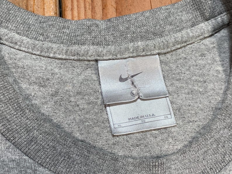 Puede incluir: Primer plano de un cuello de camiseta gris con una etiqueta Nike. La etiqueta es blanca con un logotipo Nike gris y el texto "MADE IN U.S.A.". La camiseta es de color gris liso.