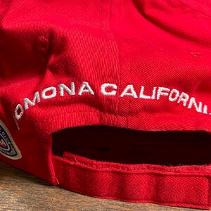 Op de afbeelding: Een felrode honkbalpet met wit geborduurde tekst die POMONA CALIFORNIA leest boven de opening aan de achterkant. Een cirkelvormige AAA Automobile Club logopatch met blauwe, witte en rode details is op de zijkant van de pet gestikt.