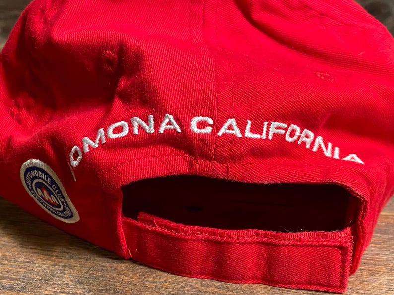 Op de afbeelding: Een felrode honkbalpet met wit geborduurde tekst die POMONA CALIFORNIA leest boven de opening aan de achterkant. Een cirkelvormige AAA Automobile Club logopatch met blauwe, witte en rode details is op de zijkant van de pet gestikt.