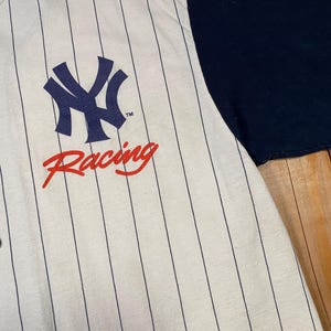 Peut inclure: Un maillot de baseball boutonn&eacute; avec une base couleur cr&egrave;me et de fines rayures verticales bleu marine. L'avant pr&eacute;sente un logo NY bleu marine au-dessus du mot Racing en &eacute;criture rouge. La chemise a une manche bleu marine et des boutons fonc&eacute;s.