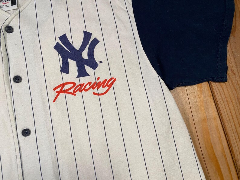 Peut inclure: Un maillot de baseball boutonn&eacute; avec une base couleur cr&egrave;me et de fines rayures verticales bleu marine. L'avant pr&eacute;sente un logo NY bleu marine au-dessus du mot Racing en &eacute;criture rouge. La chemise a une manche bleu marine et des boutons fonc&eacute;s.