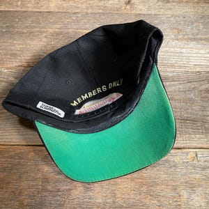 May include: Black baseball cap with a green brim. The underside of the cap features the text "MEMBERS ONLY" in gold lettering. A small white tag reads "TORONTO ELIMINATOR". The cap is set against a wooden background.
