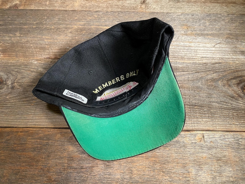 May include: Black baseball cap with a green brim. The underside of the cap features the text "MEMBERS ONLY" in gold lettering. A small white tag reads "TORONTO ELIMINATOR". The cap is set against a wooden background.