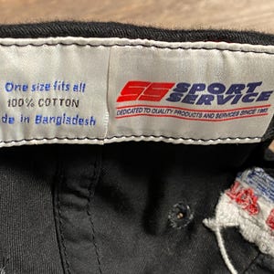 May include: Black baseball cap with a white tag that reads "One size fits all, 100% Cotton, Made in Bangladesh." The tag also features the Sport Service logo and the text "Dedicated to Quality Products and Services Since 1983."