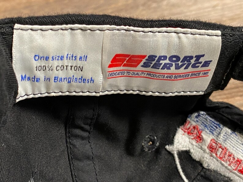 May include: Black baseball cap with a white tag that reads "One size fits all, 100% Cotton, Made in Bangladesh." The tag also features the Sport Service logo and the text "Dedicated to Quality Products and Services Since 1983."