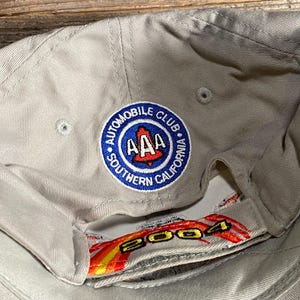 May include: A beige baseball cap featuring the embroidered Automobile Club of Southern California emblem. The emblem is a blue circle with "AAA" and a red California outline. The underside displays the year "2004" in red and yellow.