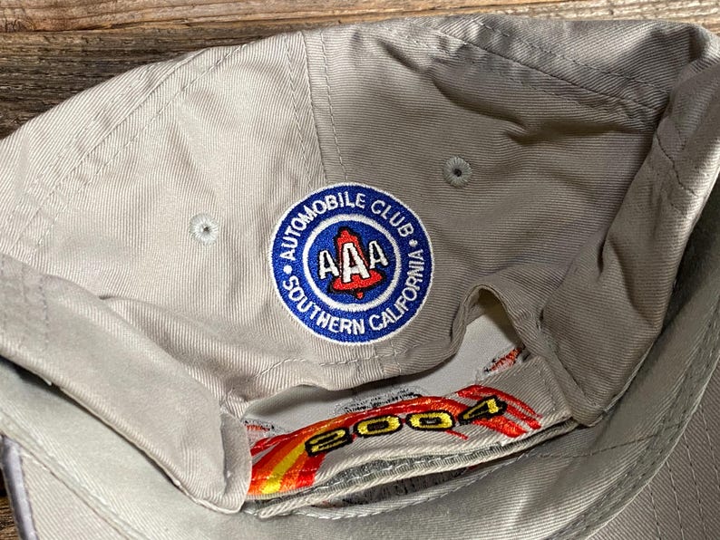 May include: A beige baseball cap featuring the embroidered Automobile Club of Southern California emblem. The emblem is a blue circle with "AAA" and a red California outline. The underside displays the year "2004" in red and yellow.