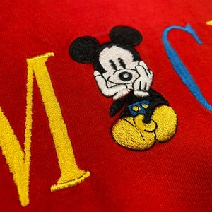 May include: A red fabric garment featuring an embroidered Mickey Mouse character positioned between large, stitched letters. The character has a white face, black ears, blue shorts, and yellow shoes. The letters are yellow and blue.