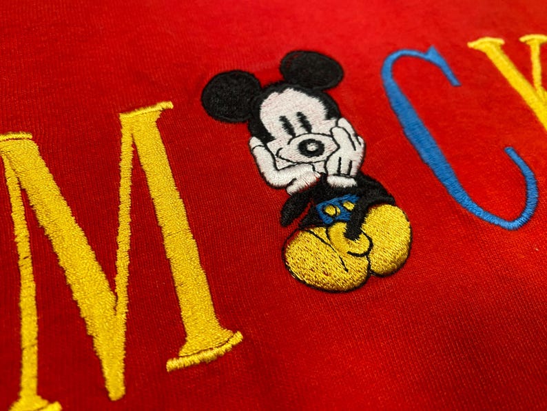May include: A red fabric garment featuring an embroidered Mickey Mouse character positioned between large, stitched letters. The character has a white face, black ears, blue shorts, and yellow shoes. The letters are yellow and blue.