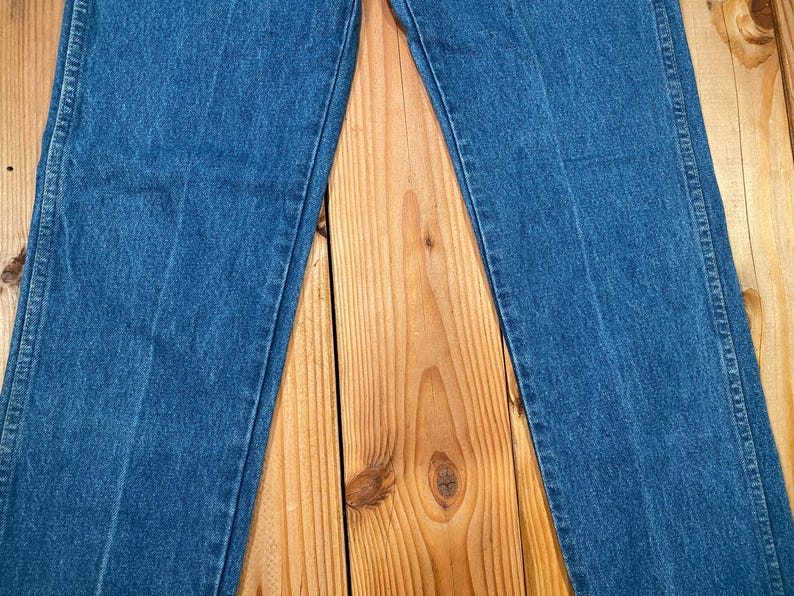 May include: Close-up of blue denim jeans. The jeans are laid flat on a wooden surface, showcasing the texture and stitching details. The denim has a classic, medium-wash appearance.