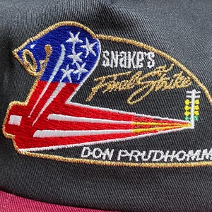 May include: Black baseball cap with a maroon brim. The embroidered patch features a stylized American flag design, the text "SNAKE'S Final Strike" and "DON PRUDHOMME" in gold. A cap for fans of drag racing.