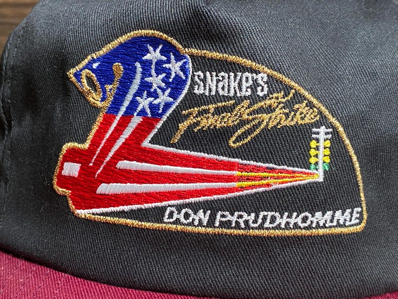 May include: Black baseball cap with a maroon brim. The embroidered patch features a stylized American flag design, the text "SNAKE'S Final Strike" and "DON PRUDHOMME" in gold. A cap for fans of drag racing.