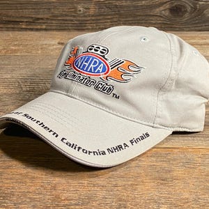 May include: A light grey baseball cap featuring the NHRA Eliminator Club logo in red, orange, and blue. The text "NHRA Finals" is on the brim. The cap is on a wooden surface.