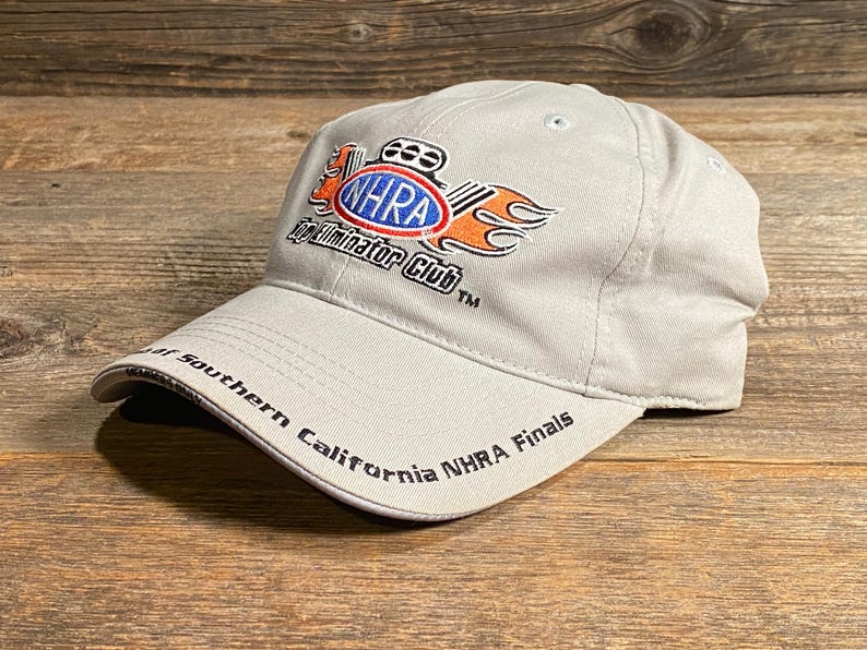 May include: A light grey baseball cap featuring the NHRA Eliminator Club logo in red, orange, and blue. The text "NHRA Finals" is on the brim. The cap is on a wooden surface.