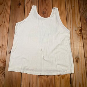 Pu&ograve; includere: Una canotta bianca con scollo tondo e un aspetto leggermente usurato. Il top senza maniche &egrave; realizzato in un tessuto morbido e leggero, adatto per l'abbigliamento casual. Il capo &egrave; appoggiato su una superficie di legno.