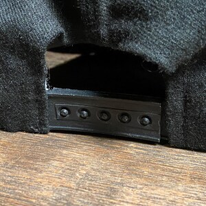May include: Close-up of the back of a black baseball cap. The cap has a black plastic snap closure and is made of a dark, textured fabric. The cap is resting on a wooden surface.