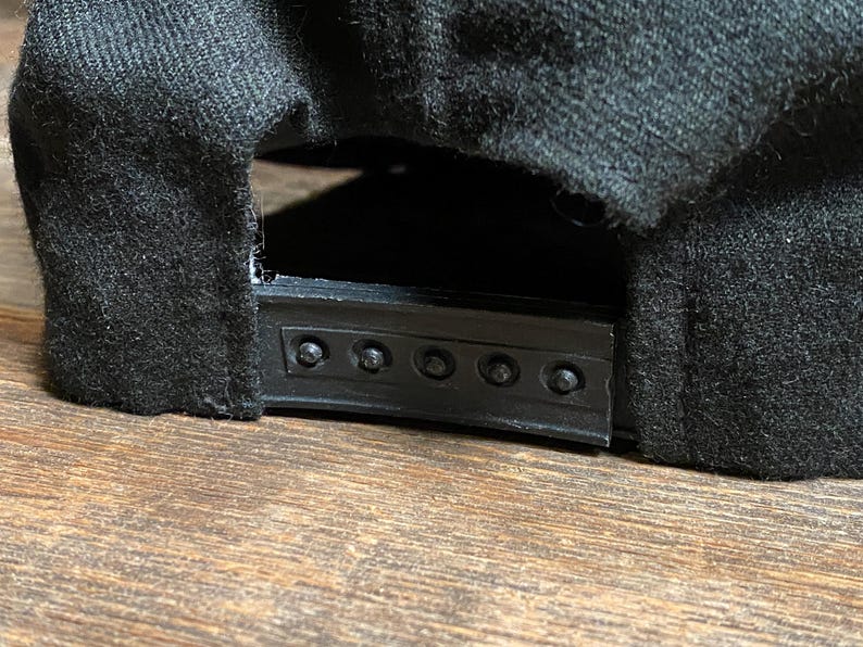 May include: Close-up of the back of a black baseball cap. The cap has a black plastic snap closure and is made of a dark, textured fabric. The cap is resting on a wooden surface.