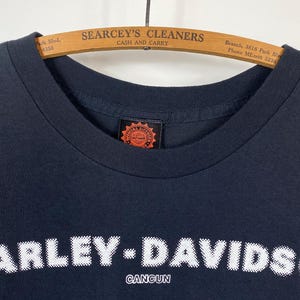 May include: Black Harley-Davidson Cancun t-shirt with white text. The Harley-Davidson logo is prominently displayed across the chest. The shirt is hanging on a wooden hanger with the text "SEARCEY'S CLEANERS CASH AND CARRY".