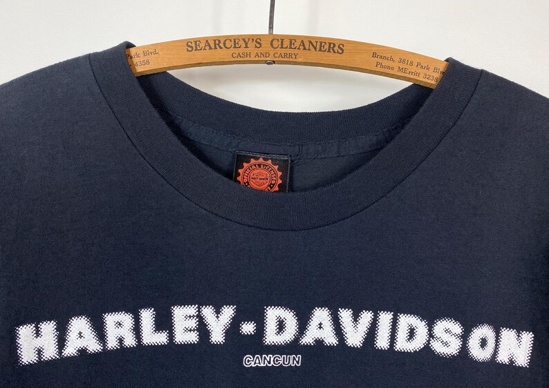 May include: Black Harley-Davidson Cancun t-shirt with white text. The Harley-Davidson logo is prominently displayed across the chest. The shirt is hanging on a wooden hanger with the text "SEARCEY'S CLEANERS CASH AND CARRY".