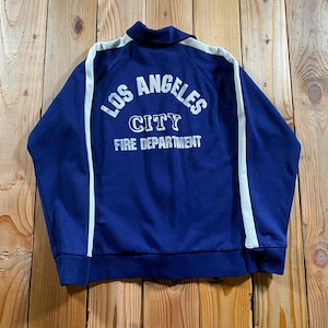 Peut inclure: Une veste zippée bleu foncé avec des bandes blanches le long des manches et un col. Le dos présente un texte imprimé blanc indiquant LOS ANGELES CITY FIRE DEPARTMENT dans une disposition empilée avec une ligne supérieure incurvée.