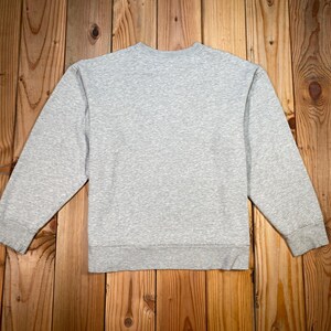 Puede incluir: Una sudadera gris jaspeado con cuello redondo y mangas largas. La sudadera est&aacute; hecha de un material suave y tiene un dobladillo y pu&ntilde;os acanalados. La prenda se exhibe sobre una superficie de madera.