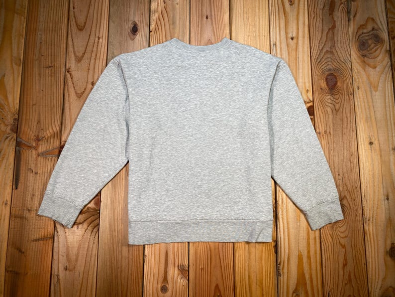 Puede incluir: Una sudadera gris jaspeado con cuello redondo y mangas largas. La sudadera est&aacute; hecha de un material suave y tiene un dobladillo y pu&ntilde;os acanalados. La prenda se exhibe sobre una superficie de madera.