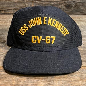 May include: A navy blue baseball cap with a flat brim. The cap features a patch with the text "USS JOHN F. KENNEDY CV-67" embroidered in gold. The cap is made of a textured fabric.