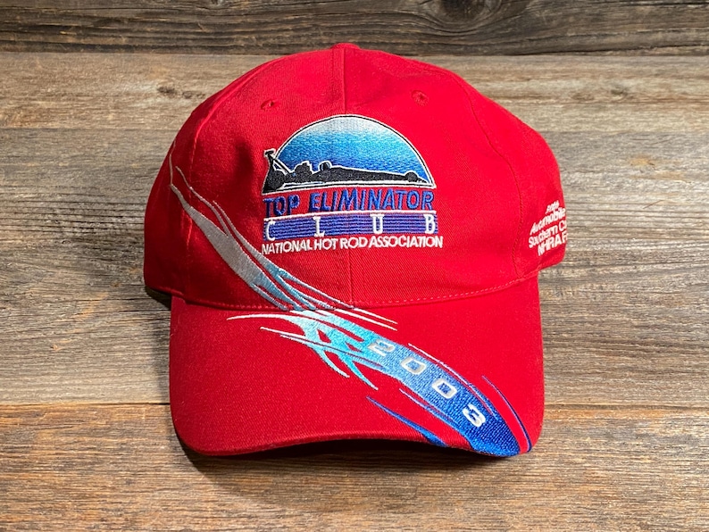 Op de afbeelding: Een felrode baseballpet met het geborduurde logo van de Top Eliminator Club National Hot Rod Association. Het ontwerp bevat een blauwe gradi&euml;nt halve cirkel, een zwart silhouet van een raceauto en blauwe tribale graphics met 2003.