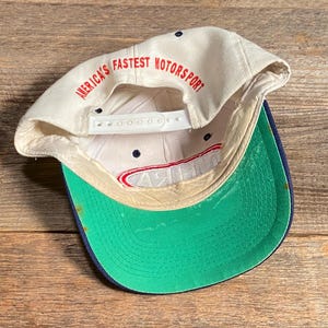 Op de afbeelding: Een vintage baseballpet met een witte kroon en een groene klep. De tekst "AMERICA'S FASTEST MOTORSPORT" is in het rood aan de binnenkant van de pet gedrukt. De pet heeft een snapback sluiting en een marineblauwe rand.