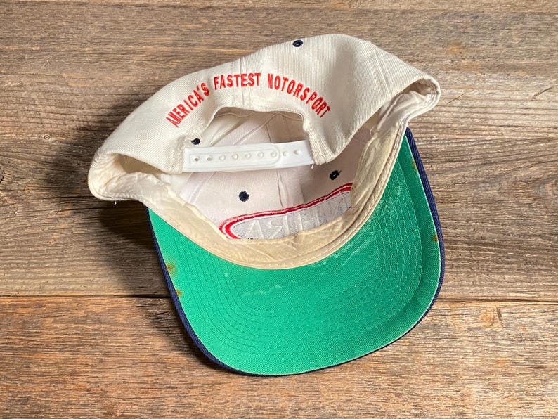 Op de afbeelding: Een vintage baseballpet met een witte kroon en een groene klep. De tekst "AMERICA'S FASTEST MOTORSPORT" is in het rood aan de binnenkant van de pet gedrukt. De pet heeft een snapback sluiting en een marineblauwe rand.