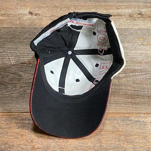 May include: Black baseball cap with a curved brim and red trim. The interior is white with black accents and red text. The cap is made of a durable fabric and features an adjustable strap.