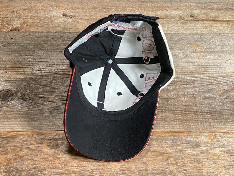 May include: Black baseball cap with a curved brim and red trim. The interior is white with black accents and red text. The cap is made of a durable fabric and features an adjustable strap.